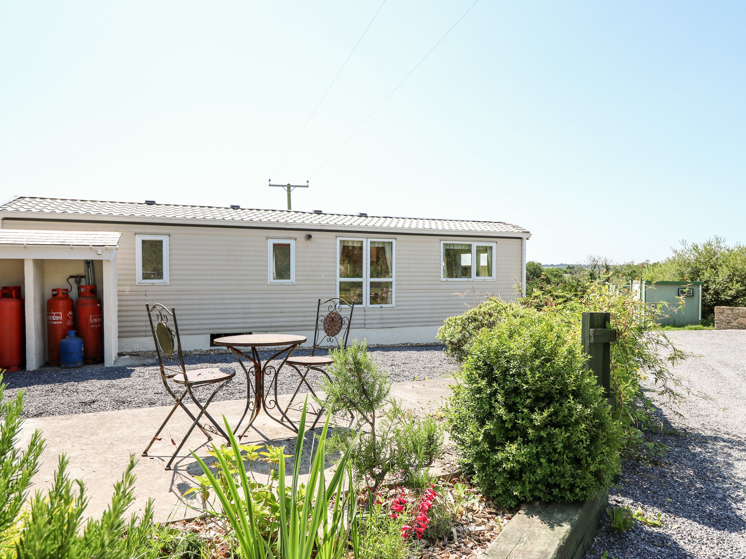 A mobile home with a patio area featuring a table and chairs, surrounded by greenery and a gravel driveway.
