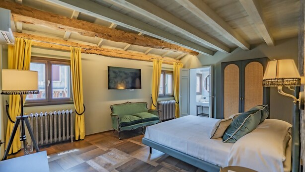 A hotel room with a large bed, a green armchair, a wooden ceiling, and a view of the outside.