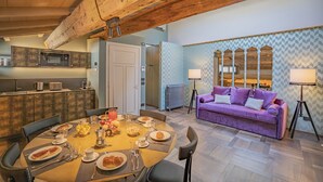 A cozy dining area with a wooden table set for breakfast, a purple sofa with lamps, and a kitchen with a microwave and toaster.