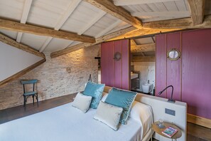 A bedroom with a bed, pillows, a chair, a small table with a magazine, and a mirror.