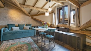 A loft-style room with exposed wooden beams, a blue sofa, a dining table with chairs, and a view of a building through a window. - Corte Rubbi 11 Secret Terrace Apartment (Venice)
