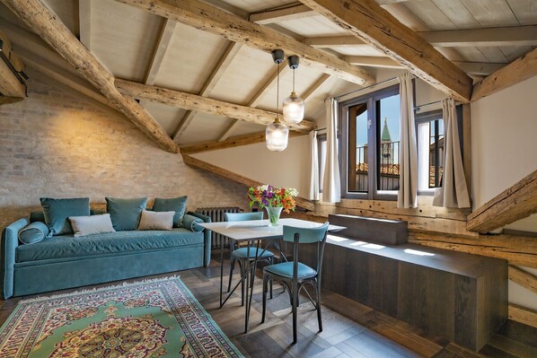 A loft-style room with exposed wooden beams, a blue sofa, a dining table with chairs, and a view of a building through a window.