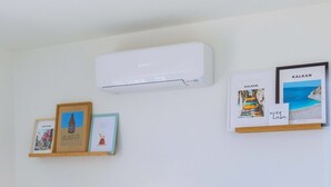 Ein Hotelzimmer mit zwei Betten, einem Nachttisch mit Lampe und Wand-Klimaanlage. An der Wand hängen gerahmte Bilder und ein Regal mit zusätzlichen Gegenständen.