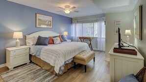 Ein Schlafzimmer mit einem Bett, Nachttischen, einem Deckenventilator, einem Fenster mit Jalousien und einem Schreibtisch mit Lampe.