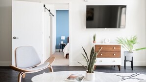 Un salón moderno con un televisor de pantalla plana, un sofá blanco, una mesa de centro con una revista, un aparador de madera y una planta en maceta.