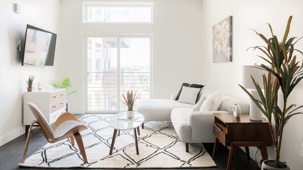 Una sala de estar moderna con un sofá blanco, una mesa de centro, un televisor montado en la pared y un amplio ventanal que proporciona luz natural.