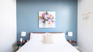 A bedroom with a bed, two bedside tables, a wall-mounted painting, and a macrame wall hanging.