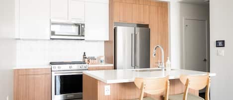 A modern kitchen with white cabinetry, a stainless steel refrigerator, a built-in oven, and a small island with two wooden chairs.