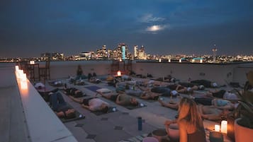Une séance de yoga sur un toit, où les participants sont allongés sur des tapis, entourés de bougies, et d'où l'on peut admirer le panorama d'une ville au crépuscule.