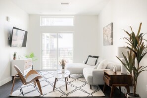 Ein modernes Wohnzimmer mit einer Couch, einem Couchtisch, einem Sessel, einem an der Wand montierten Fernseher und einem großen Fenster.