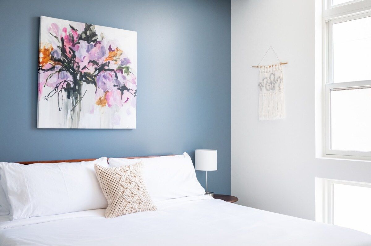 Un dormitorio con cama, ropa de cama blanca, un cuadro colgado en la pared, una lámpara de noche y un colgador de macramé.