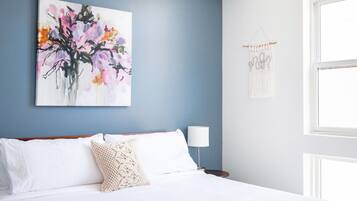 Un dormitorio con cama, ropa de cama blanca, un cuadro colgado en la pared, una lámpara de noche y un colgador de macramé.