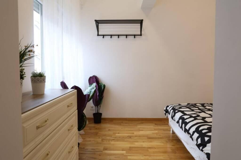 A bedroom with a wooden floor, a bed with a patterned bedspread, a dresser with drawers, a wall-mounted shelf, and a potted plant.
