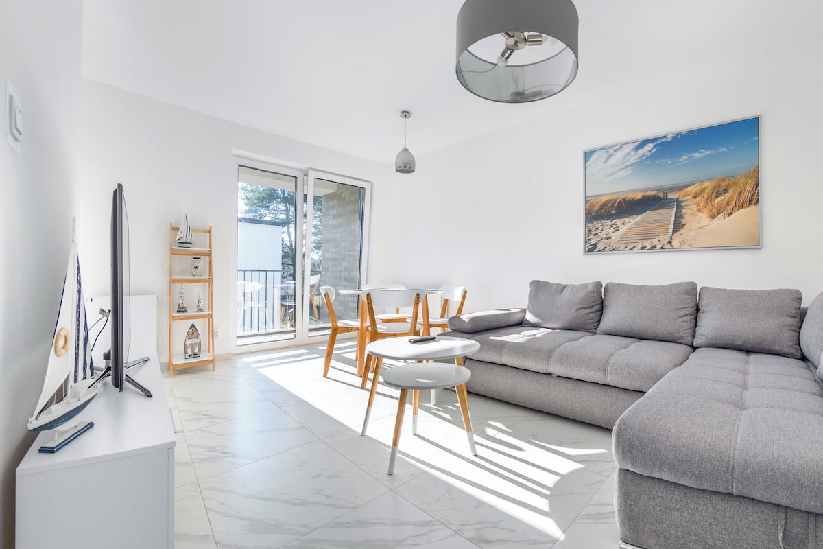 A modern living room with a grey sofa, wooden chairs, a white coffee table, a TV, and a bookshelf.