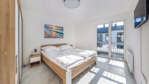 A bedroom with a wooden bed, white bedding, a painting of a sunset, a nightstand, a TV, and a balcony with a view of buildings.