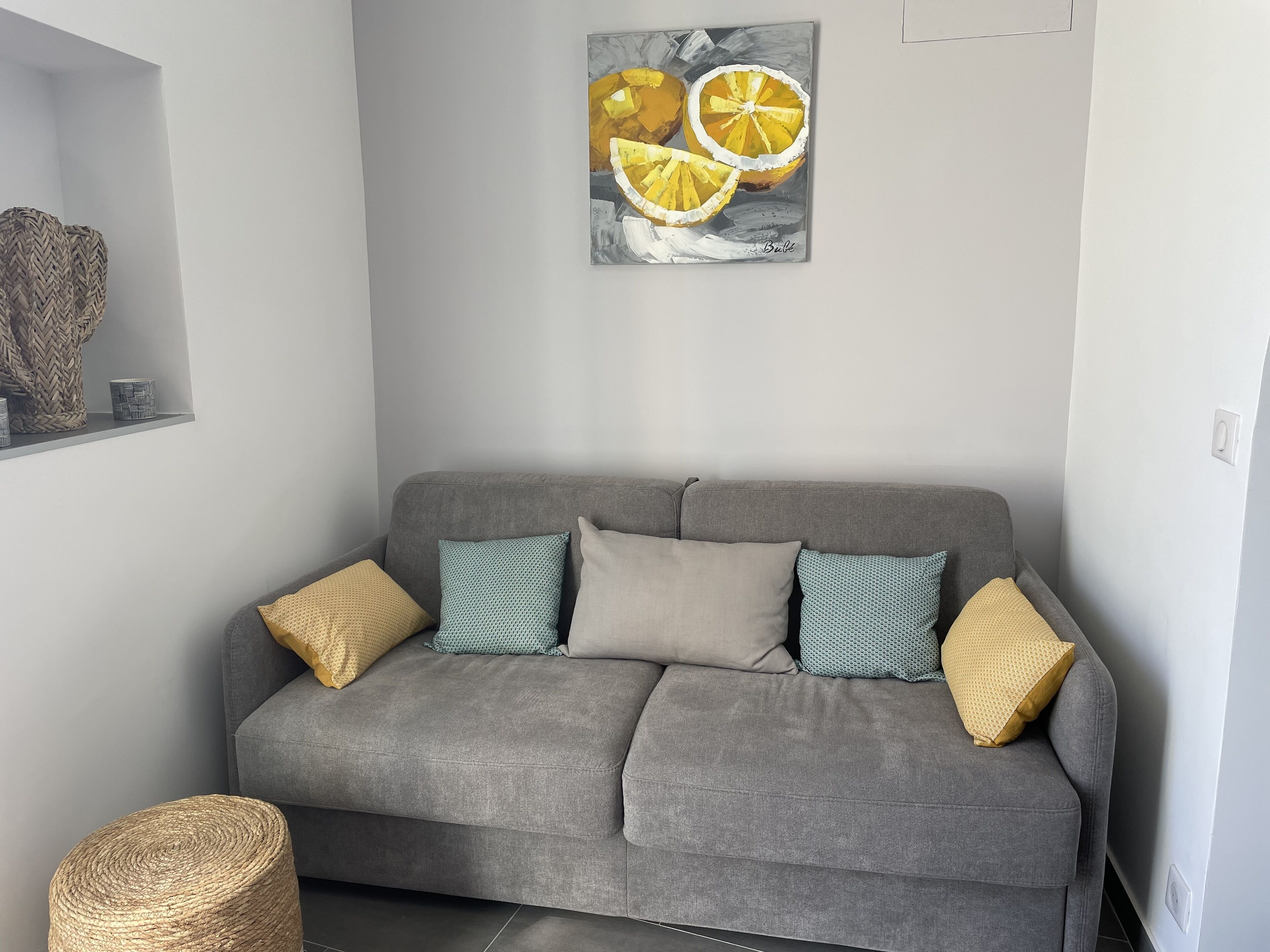 A grey sofa with patterned cushions, a woven basket, and a painting of lemons on the wall.