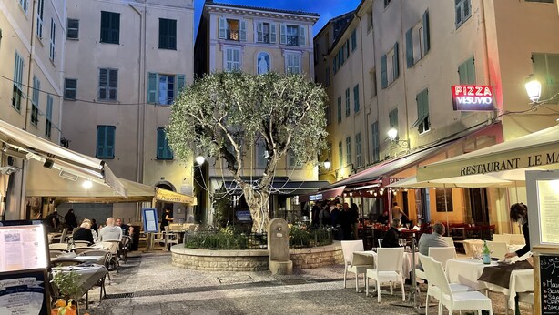Een terras met tafels en stoelen, omringd door gebouwen met borden voor een pizzeria en een patisserie.