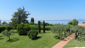 A well-maintained garden with a variety of trees and shrubs, a clear blue sky, and a view of a body of water in the distance. - Regarda - Blue View 2 with lake view, 2 bedrooms, pool (Bardolino)