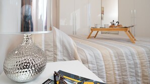 Une chambre d’hôtel bien rangée, avec une couvre-lit à rayures, une table avec une lampe et, au fond, un coin repas.