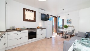 Une cuisine moderne avec des meubles blancs, une cuisinière à gaz et un four encastré. On y trouve un coin repas avec une table et des chaises, ainsi qu’un canapé gris.