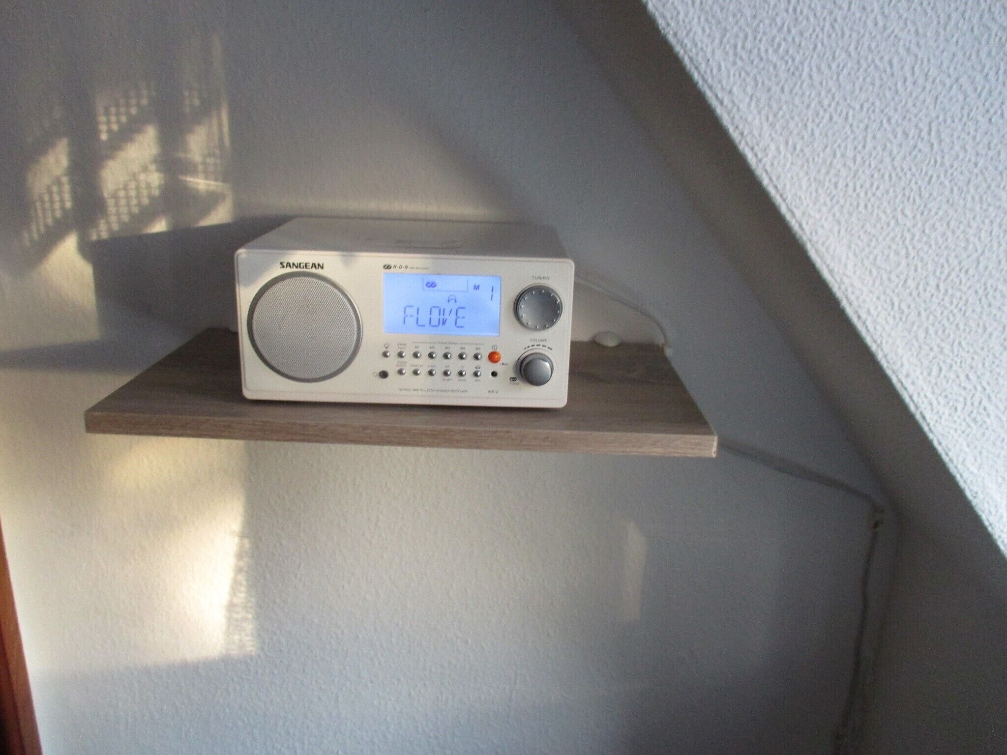 Een Sangean radio op een houten plank tegen een muur.