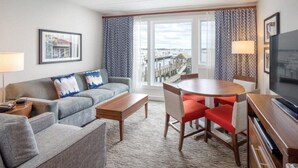 Quarto de hotel com sofá cinzento, mesa de centro de madeira, mesa de jantar com cadeiras vermelhas, televisão e vista para a marina através de grandes janelas.