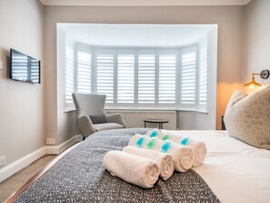 Habitación de hotel con una cama grande, una silla, una mesita y un ventanal con contraventanas blancas.