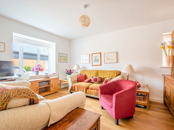 A cozy living room with a sofa, armchair, and a wooden coffee table. There is a window with a view of the outside, a TV, and a lamp on a side table.