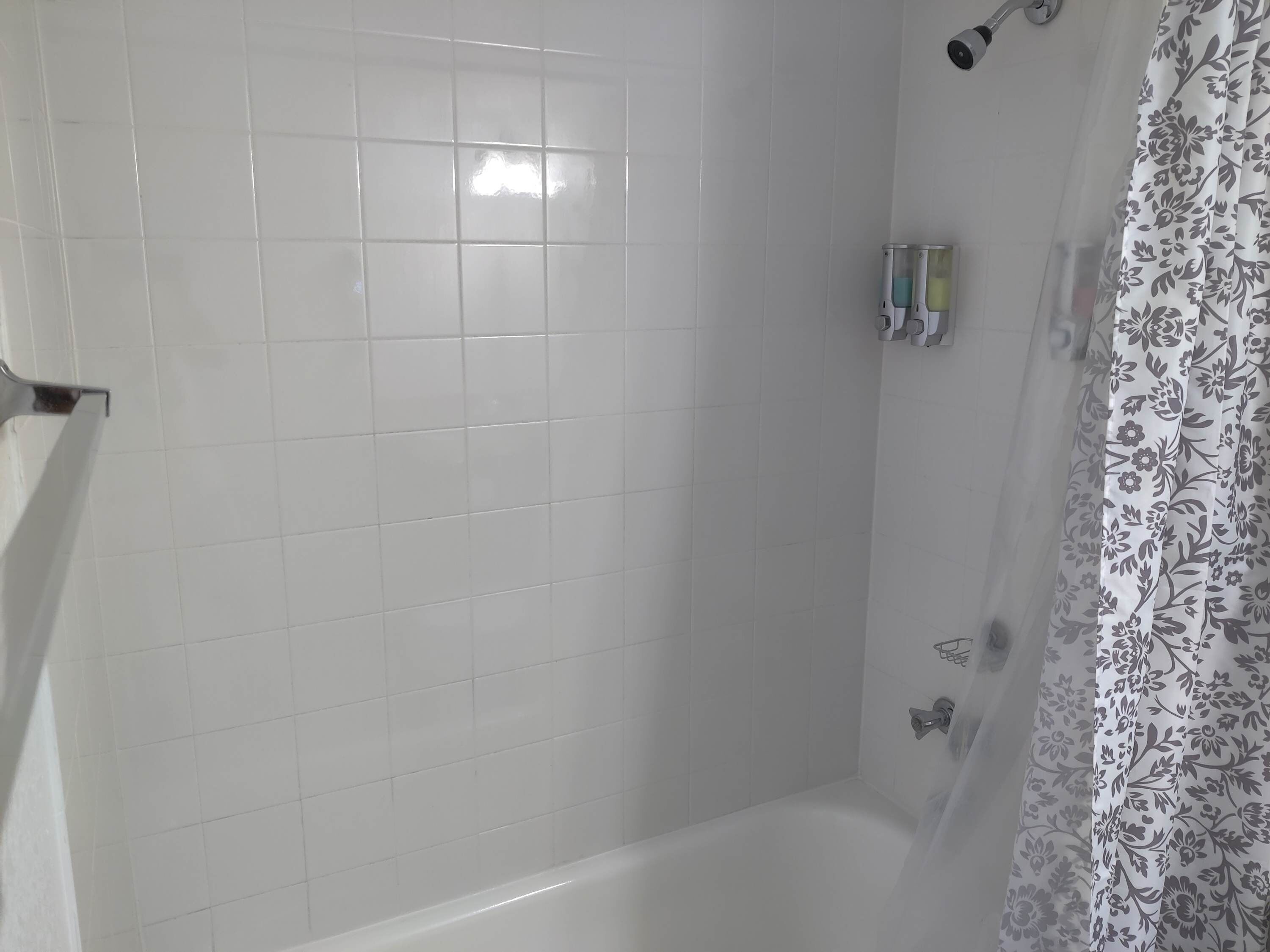 A bathroom with white tiled walls, a bathtub, a shower, and a floral-patterned shower curtain.