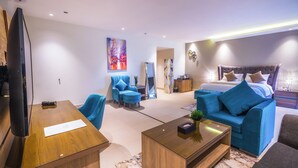 A modern hotel room with a large bed, a seating area with a blue sofa and armchairs, a wooden coffee table, and a flat-screen TV. - THE ADDRESS PALACE HOTEL & APARTMENT (Dammam)