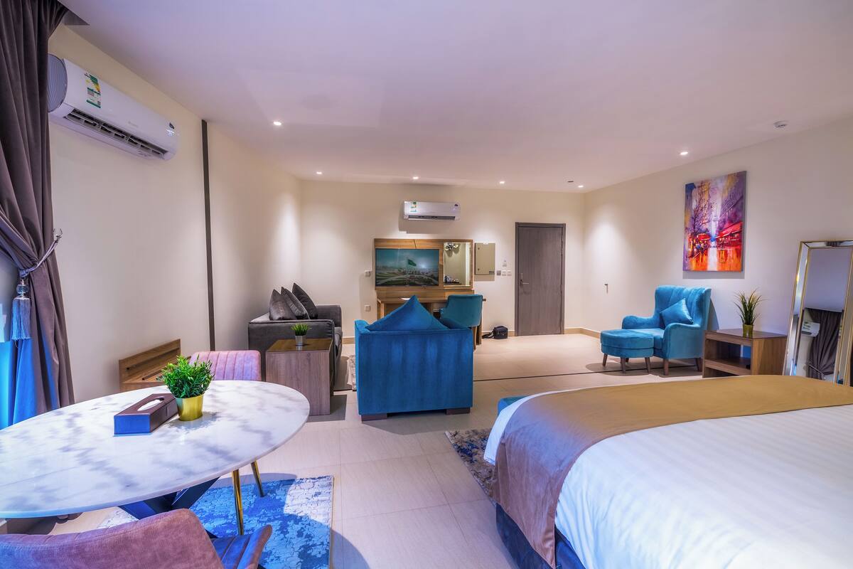 A modern hotel room with a bed, two blue armchairs, a small table, a desk with a chair, and an air conditioning unit.