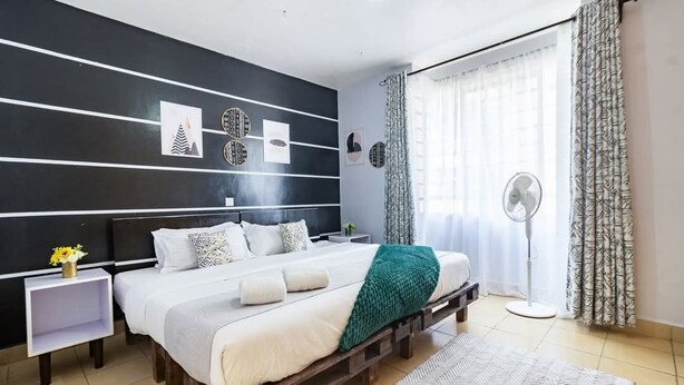 A modern bedroom with a bed, bedside tables, a fan, and a window with curtains. - Hirwado Homes - QUIET/Secure/15 Mins to JKIA /SGR (Mlolongo)