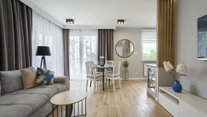 A modern living room with a grey sofa, a small round table, white chairs, a dining table set for two, and a large window with curtains. - Prywatne apartamenty w Sun & Snow Porta Mare (Kolobrzeg)