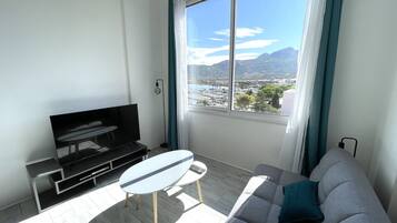 A modern living room with a TV, sofa, and a small table. A large window offers a view of a city and mountains.