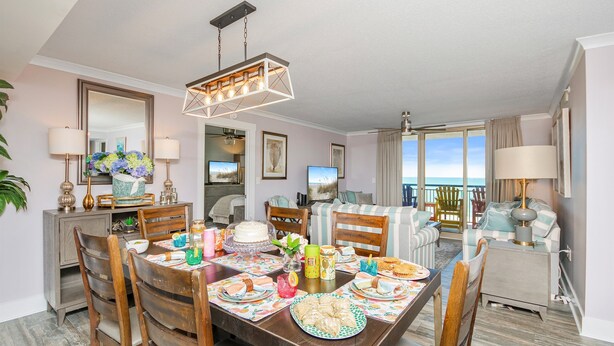 A dining area with a wooden table set for a meal, a chandelier, and a view of the ocean through large windows. - Oceanviews-Steps 2 Beach-Stunning-MultiBalconies-Gated Community-Pool/Hottub (Myrtle Beach)