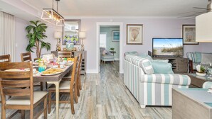 A dining area with a wooden table set for a meal, a sofa with striped cushions, and a television in the living room. - Oceanviews-Steps 2 Beach-Stunning-MultiBalconies-Gated Community-Pool/Hottub (Myrtle Beach)