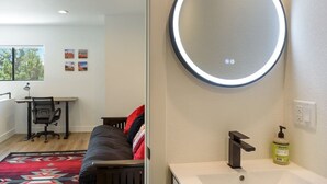 Une chambre à coucher avec un lit, un bureau avec une chaise, un miroir rond et une salle de bain avec un lavabo et un flacon de savon liquide.