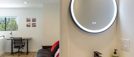 A bedroom with a bed, a desk with a chair, a round mirror, and a bathroom with a sink and a bottle of hand soap.