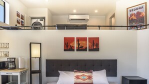 Une chambre d’hôtel comprenant un lit, une table de chevet avec une machine à café, un climatiseur fixé au mur et deux tableaux représentant des paysages désertiques.