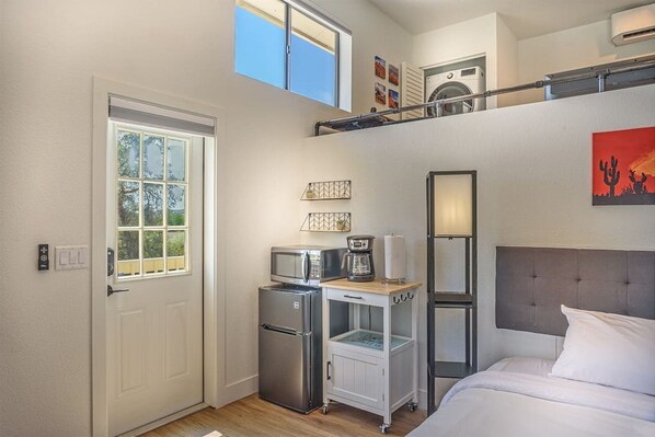 Una habitación de hotel compacta con cama, kitchenette equipada con refrigerador, microondas y cafetera, una ventana pequeña y un ventilador de techo.