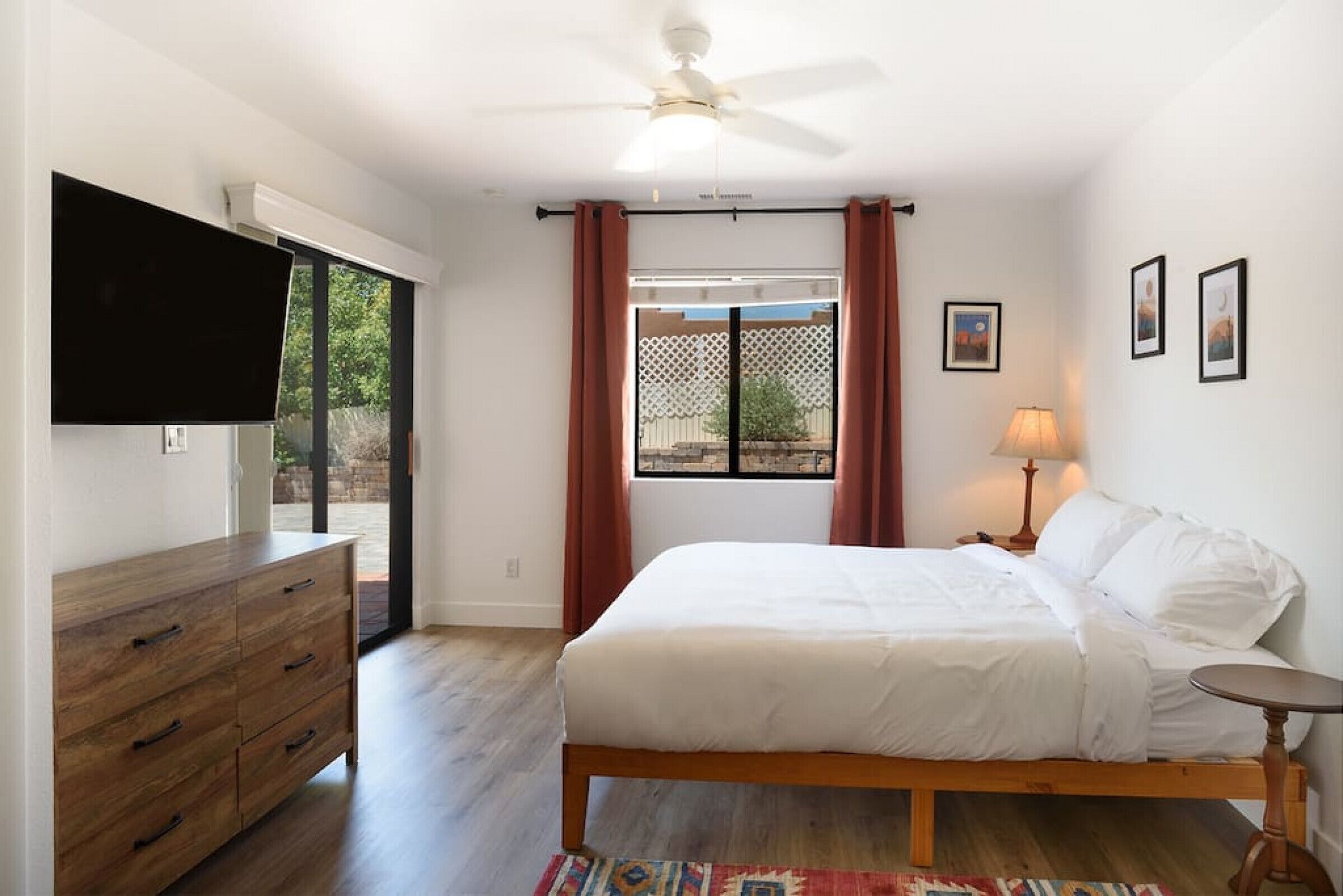 Ein Schlafzimmer mit Bett, Kommode, Deckenventilator, Fernseher, Fenster mit Vorhängen und einem kleinen Tisch mit Lampe.
