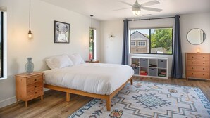 A bedroom with a bed, wooden nightstands, a ceiling fan, a window with curtains, and a view of a house outside. - Modern Zen Luxury · With Views · Centrally Located (Sedona)