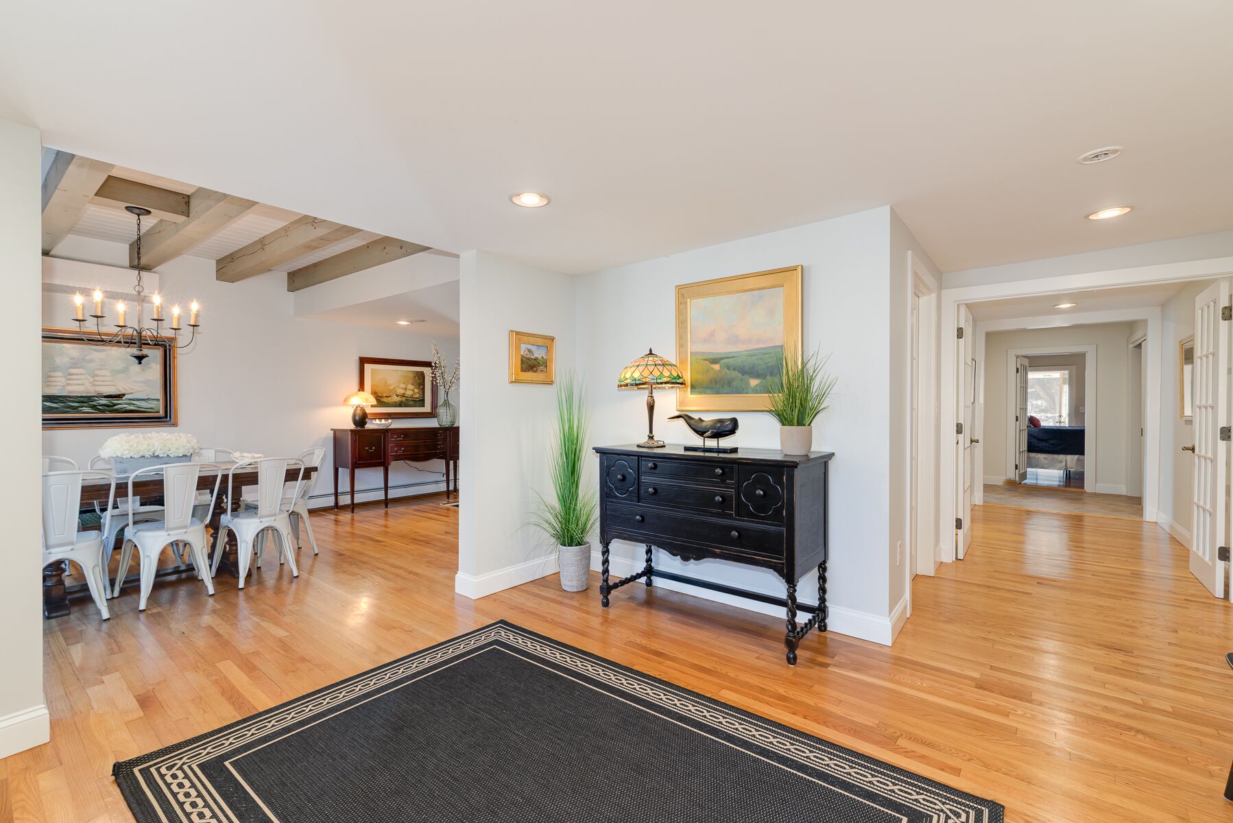 A spacious living area with hardwood flooring, a dining table with chairs, a black console table with decorative items, and a painting on the wall.