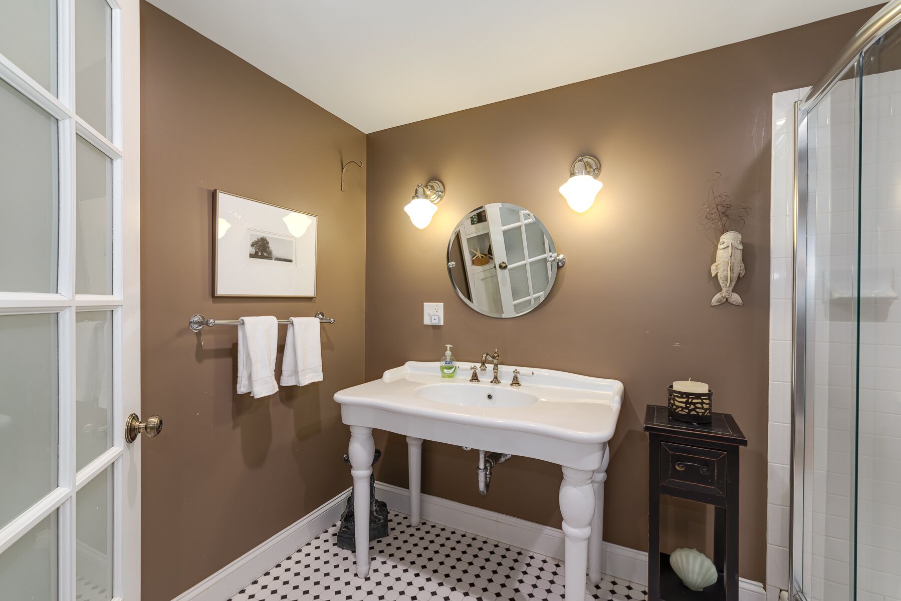 A bathroom with a white sink, two wall-mounted lights, a round mirror, a framed picture, and a decorative fish on the wall.