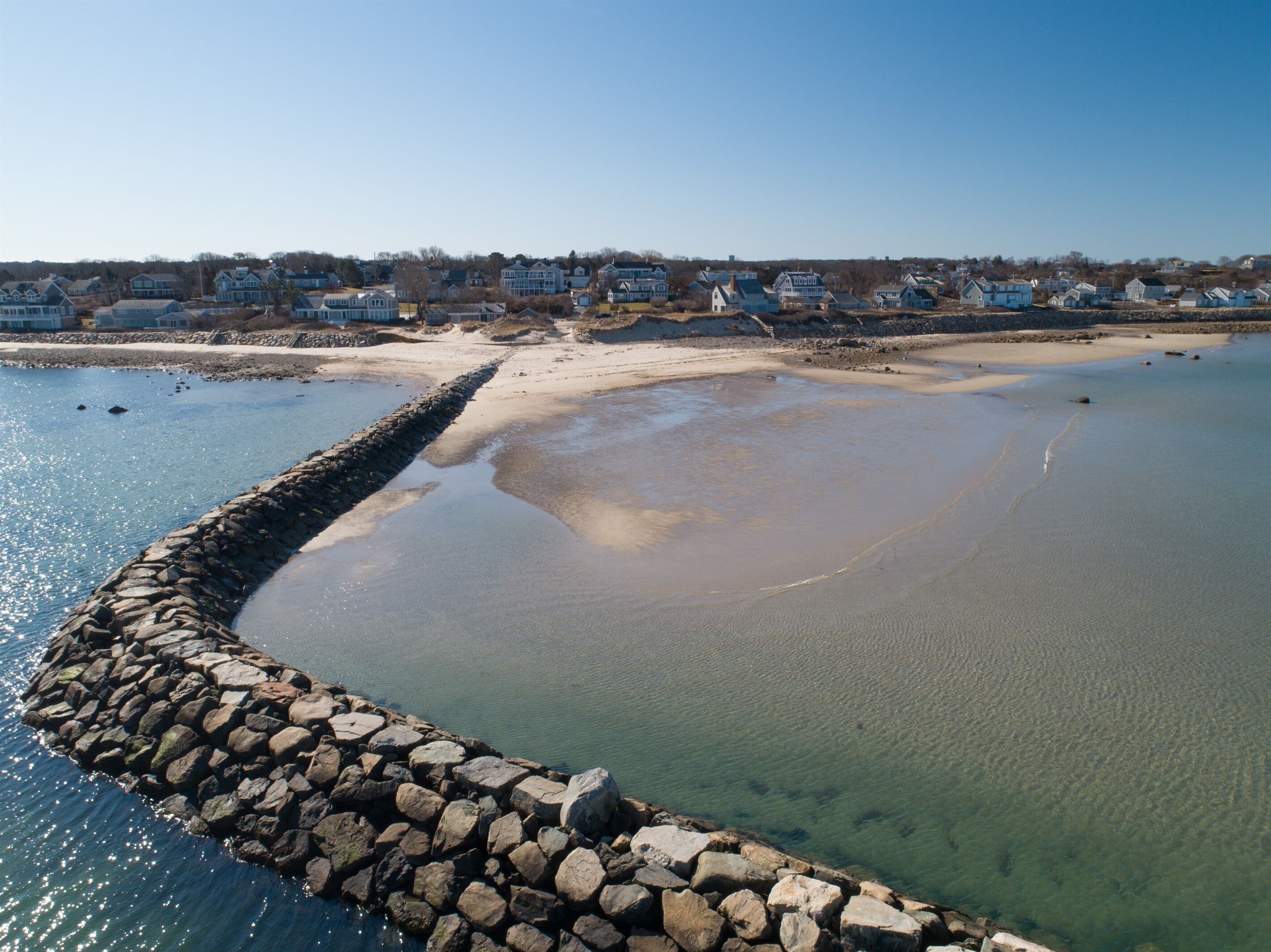 A coastal area with a rocky breakwater, calm water, and a row of houses in the background.