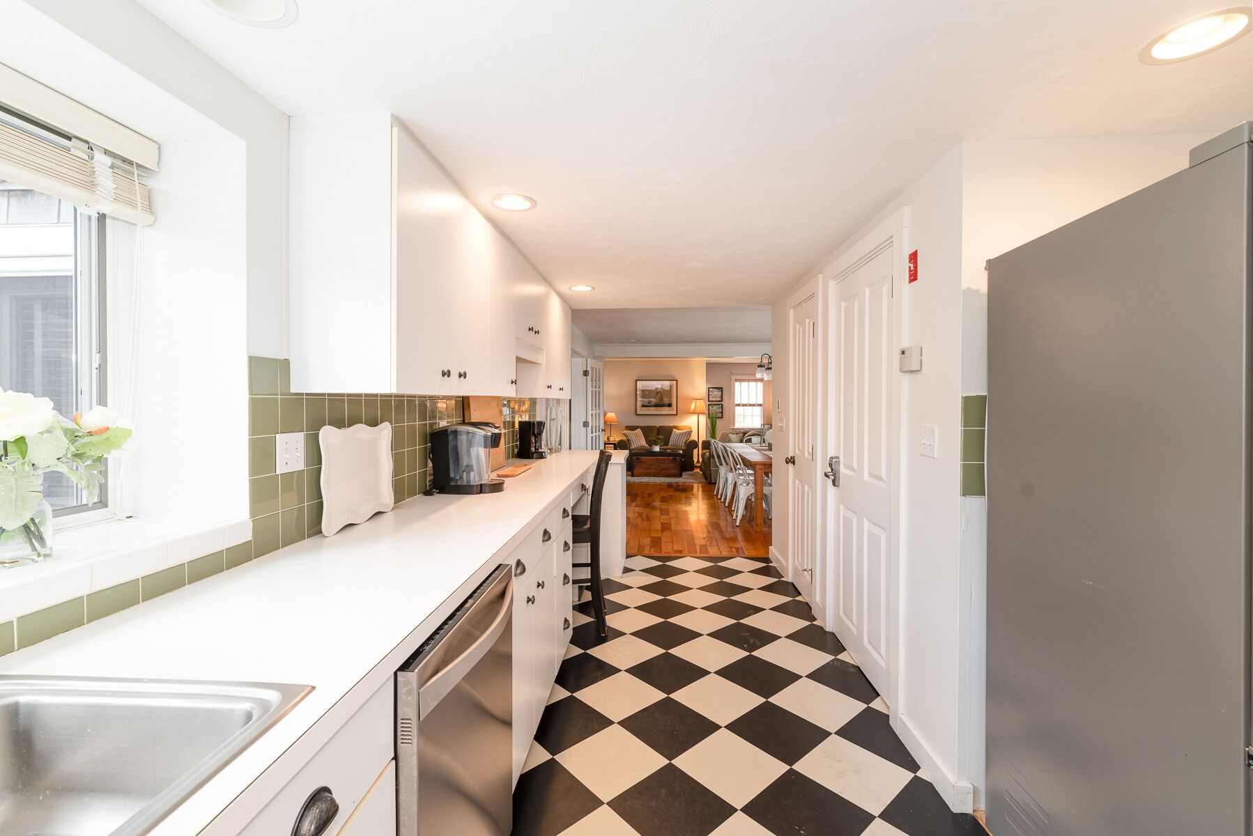 A narrow hallway with a checkered floor, leading to a kitchen area with a coffee machine and a dining table.