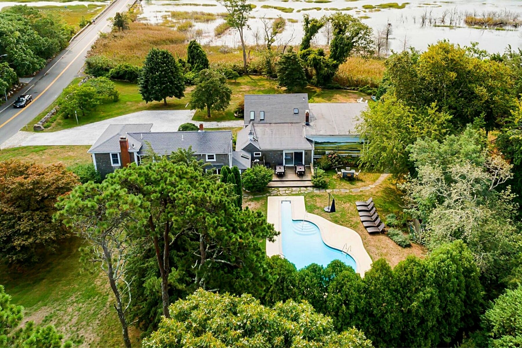 A large house with a swimming pool, surrounded by dense trees and a road in the background.