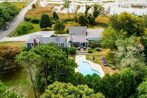 A large house with a swimming pool, surrounded by dense trees and a road in the background.