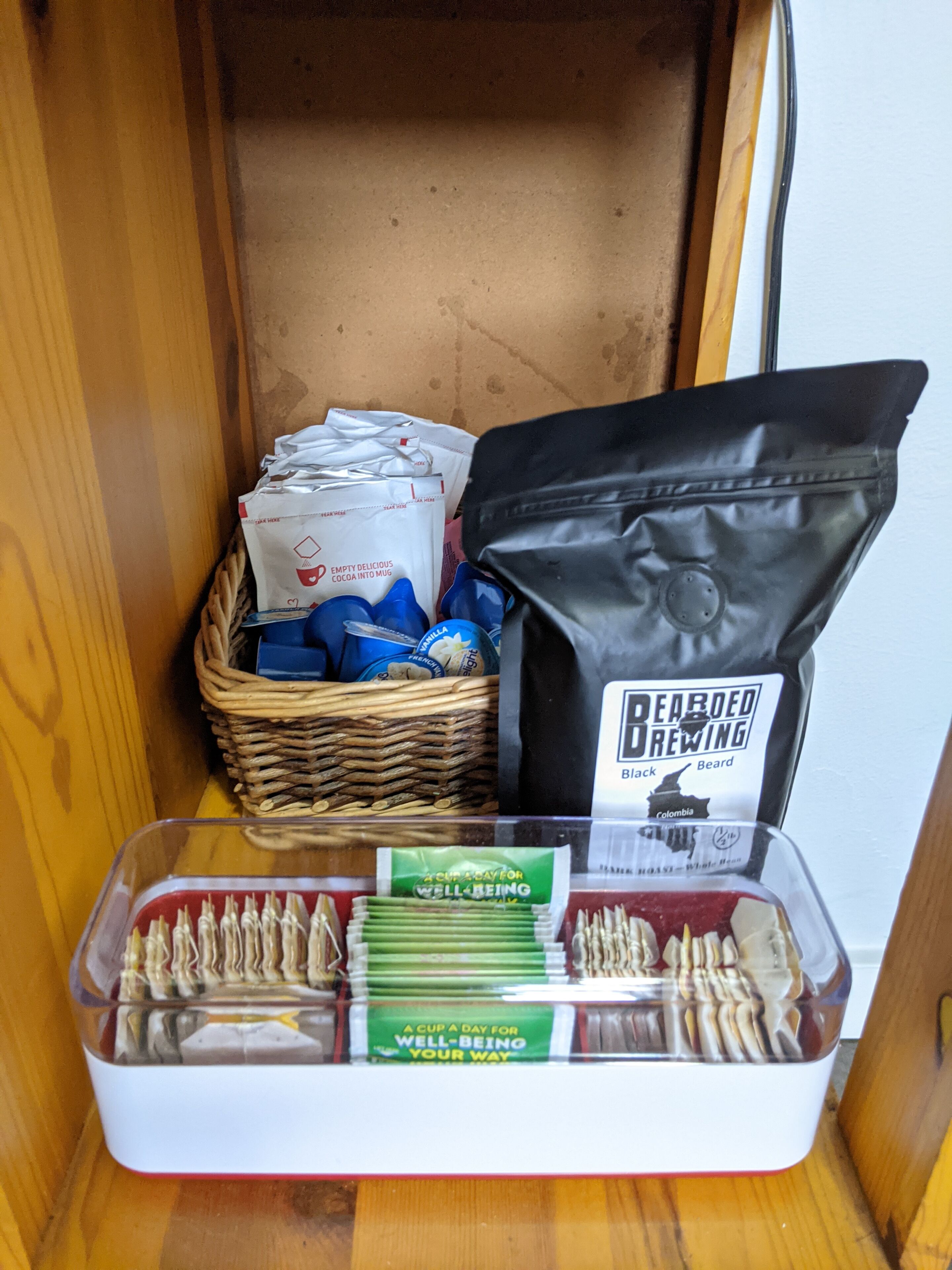 Un espace de rangement contenant un panier en osier rempli de sacs de café, un sac noir étiqueté « Bearded Brewing » et un récipient contenant des sachets de thé.