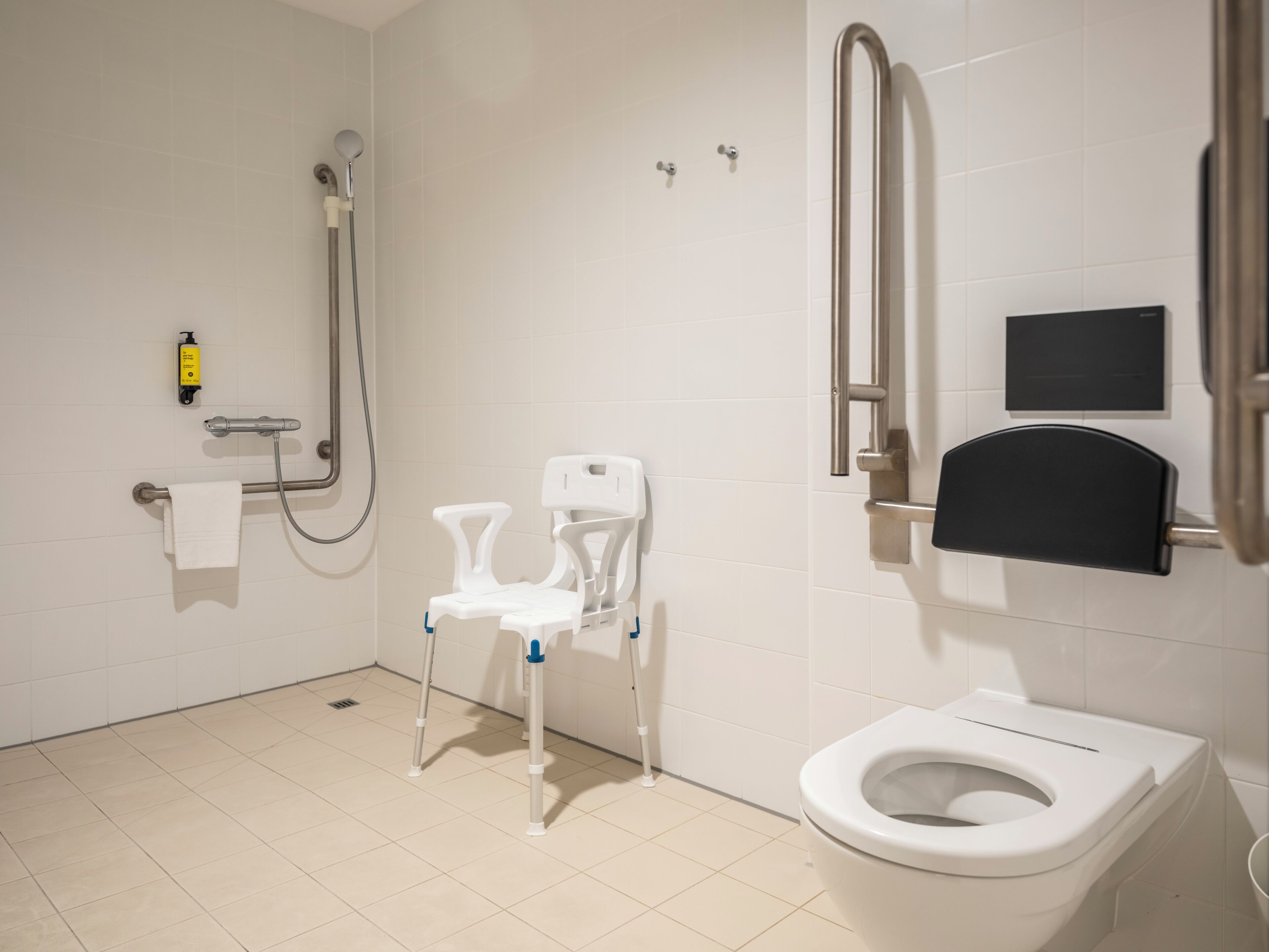 Un baño moderno con inodoro blanco, ducha con cabezal de ducha de mano, barra de apoyo y una silla blanca.